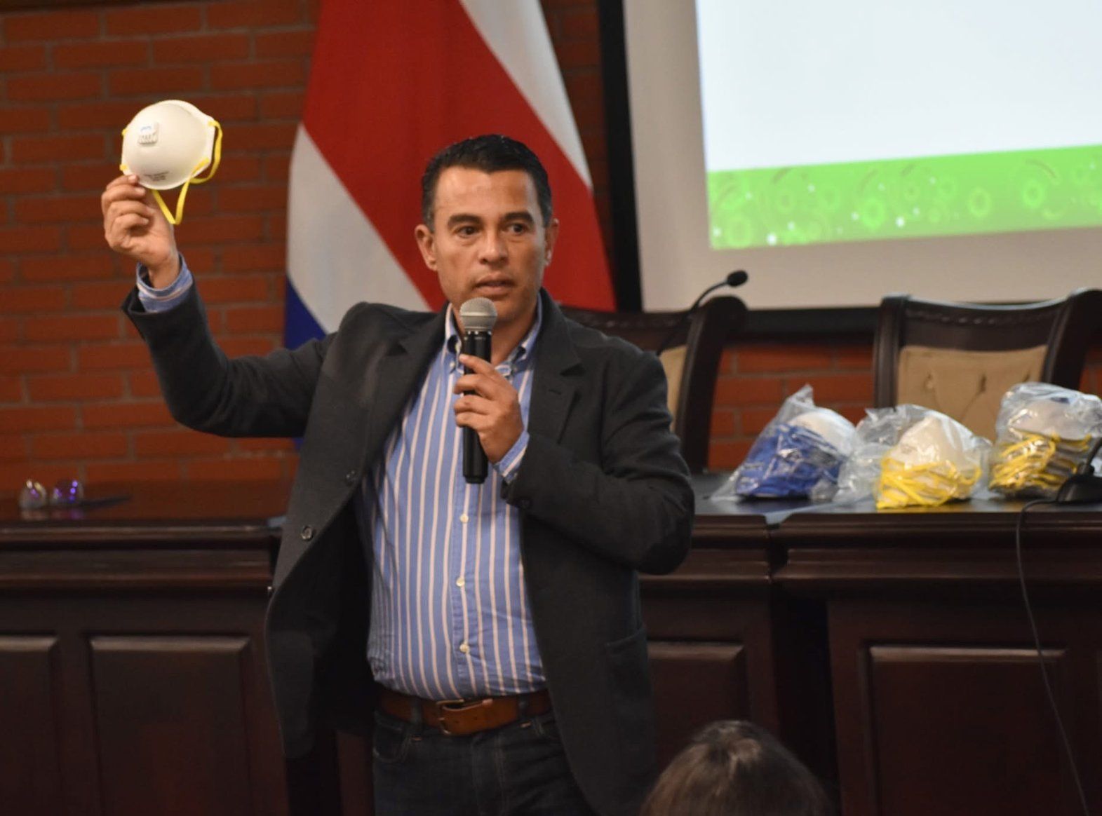Roy Sosa mostrando un respirador desechable durante una conferencia, con la bandera de Costa Rica al fondo